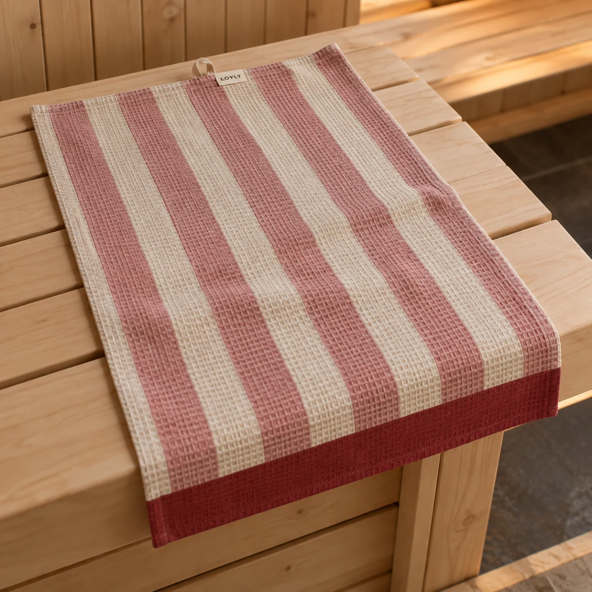 Toalha de assento Favo Goiaba, listras rosa e cru com barra vermelha, sobre um banco de sauna
